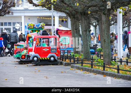 Sopot, Polen - 11.11.2024: Ein kleiner roter Spielzeugwagen in der Nähe von Bänken und Bäumen auf dem Sopot-Platz, mit Gebäuden im Hintergrund. Stockfoto