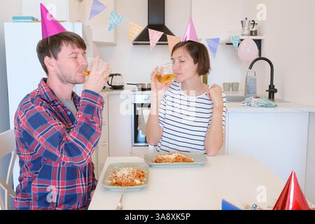 Das junge Paar feiert zu Hause gemütlich Geburtstag und genießt Kuchen und Lachen in warmer Atmosphäre. Die Familie isst Pasta und trinkt Wein zum Feiern Stockfoto