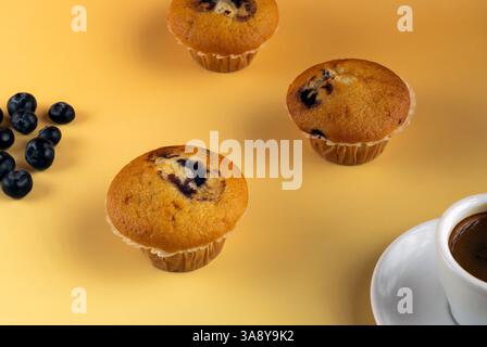 Drei Muffins mit Heidelbeeren auf einem gelben Tisch. Eine Tasse Kaffee ist neben ihnen Stockfoto