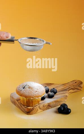 Eine Person streut Puderzucker auf einen Muffin. Der Muffin ist mit Heidelbeergeschmack geschmeckt und sitzt auf dem hölzernen Schneidebrett. Orangefarbener warmer Hintergrund Stockfoto