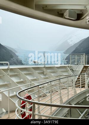 Das vordere Deck eines Kreuzfahrtschiffs bietet einen atemberaubenden Blick auf einen blauen Gletscher, eingebettet zwischen nebligen, schneebedeckten Bergen in einem abgelegenen Fjord. Stockfoto