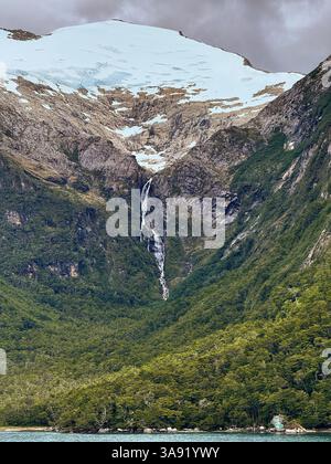 Ein schmaler Wasserfall fällt von einem Gletscher hoch in den verschneiten Bergen, schneidet durch ein bewaldetes Tal und fließt in Richtung eines türkisfarbenen Fjords darunter. Stockfoto