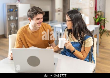 Junge multiethnische Paare, die sich lächeln und anschauen, während sie einen Laptop benutzen und Kaffee trinken, am Tisch in ihrer modernen Wohnung sitzen Stockfoto