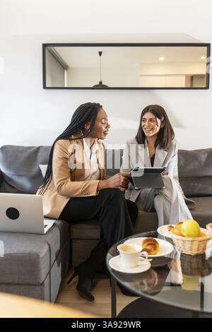 Zwei Geschäftsfrauen sitzen auf einem Sofa in einem Hotelzimmer, verwenden ein Tablet und einen Laptop während eines produktiven Meetings und genießen Kaffee und Croissants Stockfoto