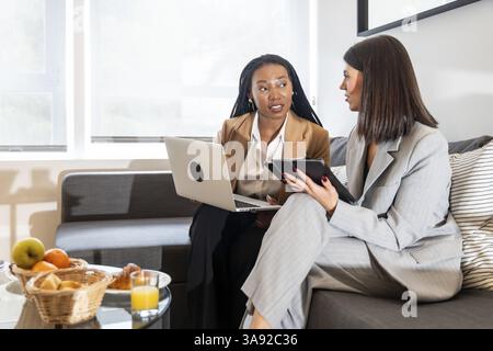 Zwei Geschäftsfrauen, die Laptop und Tablet verwenden, diskutieren während einer Geschäftsreise im Hotelzimmer über die Arbeit Stockfoto