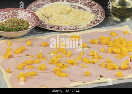 Teigboden mit Wurstscheiben, Paprika und Kräutern, umgeben von Zutaten auf Tellern Stockfoto