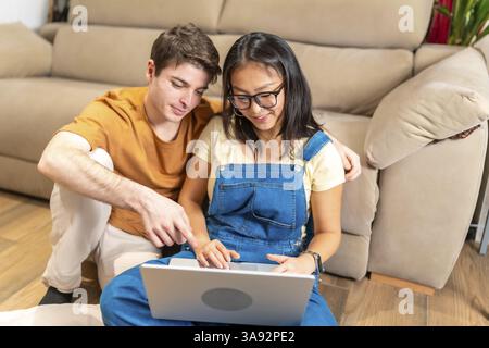 Junge multiethnische Paare sitzen bequem auf dem Boden ihres gemütlichen Wohnzimmers und nutzen einen Laptop, um gemeinsam im Internet zu surfen Stockfoto
