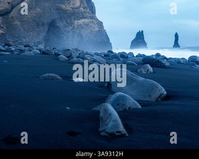 Nahaufnahme glatter vulkanischer Steine, die auf dem schwarzen Sand des Reynisfjara Beach verstreut sind, mit Basaltklippen und Reynisdrangar-Meeresstapeln, die aus dem entspringen Stockfoto
