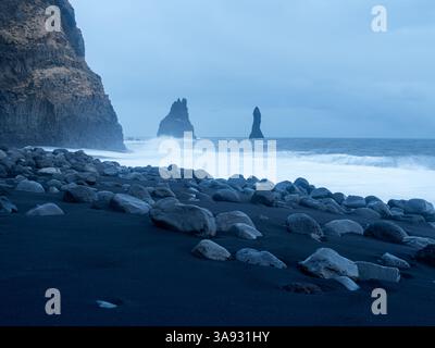 Reynisfjara Beach in der Abenddämmerung mit zerklüfteten Basaltsäulen und Reynisdrangar-Meeresstapeln. Der schwarze Sandstrand ist von glatten vulkanischen Felsen verstreut Stockfoto