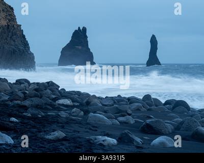 Reynisfjara Beach in der Abenddämmerung mit den berühmten Reynisdrangar-Meeresstapeln, die aus dem Ozean ragen, eingerahmt von dunklen Basaltsäulen und verstreuten vulkanischen roc Stockfoto