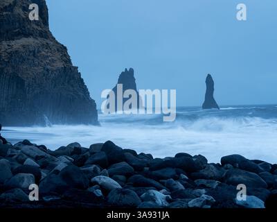 Reynisfjara Beach in der Abenddämmerung, mit den ikonischen Reynisdrangar-Meeresstapeln, die vor turbulenten Wellen stehen, eingerahmt von dunklen Basaltsäulen und verstreuten wolken Stockfoto
