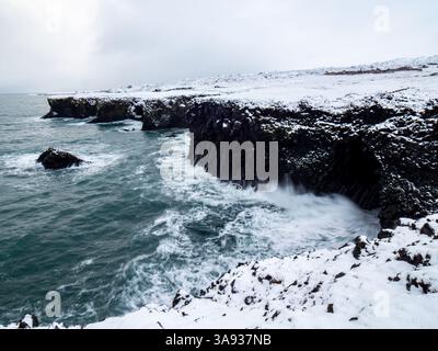 Schneebedeckter Felsbogen Gatklettur in Arnarstapi, Island. Winterwellen stürzen in die dramatische Basaltküste der Halbinsel Snæfellsnes Stockfoto