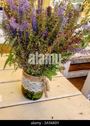 Rustikaler Blumenstrauß in einem Glasgefäß mit Spitze und Zwirn auf Vintage-weißem Holztisch in ländlicher Umgebung Stockfoto