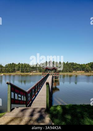 Traditioneller chinesischer Pavillon mit rotem Geländer auf einer Holzbrücke über einen See in einem friedlichen Park an einem klaren, sonnigen Tag Stockfoto