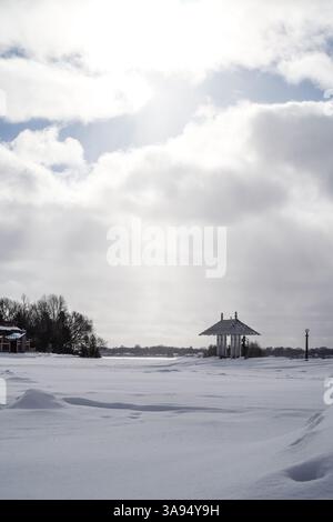 Eine verschneite Winterlandschaft in Kanada bietet einen weißen Pavillon und Bäume unter einem bewölkten Himmel. Fußabdrücke sind auf dem schneebedeckten Boden sichtbar. Stockfoto
