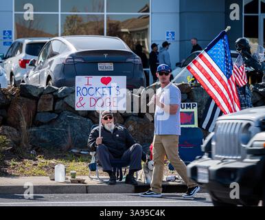 Ein Mann sitzt mit einem Schild über die Liebe zu Raketen und DOGEN, während ein anderer mit einer US-Flagge vor dem Tesla-Händlerbetrieb bei einem Takeback-Protest vorbeigeht. Stockfoto