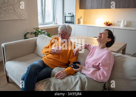 Glückliche lachende Ehemann- und Ehefrau-Paare, die gemeinsam auf einer gemütlichen Couch im Wohnzimmer Tee genießen Stockfoto
