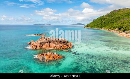 Eine ruhige Küstenszene mit zerklüfteten Felsen, die aus ruhigen smaragdgrünen Gewässern in der Nähe eines grünen Hügels auftauchen. Praslin, Seychellen. Stockfoto