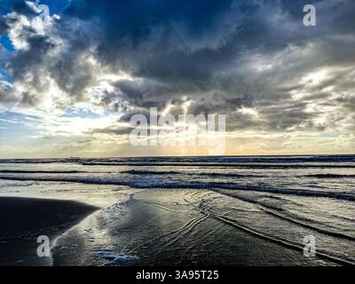 "Ein ruhiger Morgen am Meer, mit sanften Sonnenstrahlen, die durch die Wolken brechen und ein goldenes Licht über das ruhige Wasser werfen." Stockfoto