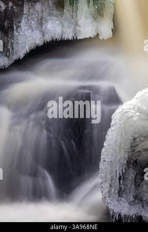 Nahaufnahme eines gefrorenen Wasserfalls mit nebligem fließendem Wasser, kurzen Eiszapfen, die von Felsen hängen, und Eisformationen, die die Steinoberfläche bedecken Stockfoto