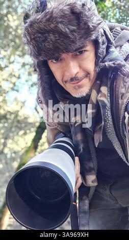 Tierfotograf mit Teleobjektiv in der Natur. Ein Mann in Tarnkleidung und Winterkleidung schaut in die Kamera, während er einen professionellen CA hält Stockfoto