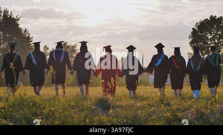 Absolventen gehen bei Sonnenuntergang zusammen auf einem Feld, halten Hände, symbolisieren Freundschaft und eine gemeinsame Zukunft Stockfoto