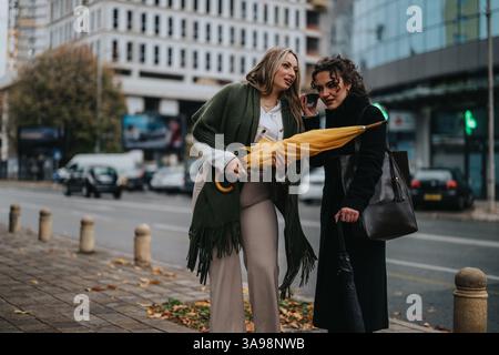 Zwei stilvolle Frauen sprechen und teilen sich einen Regenschirm auf einem Bürgersteig der Stadt Stockfoto