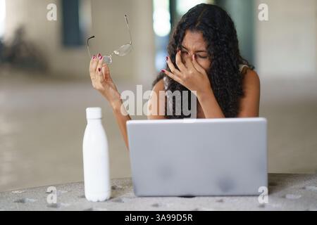 Eine frustrierte Frau, die Schwierigkeiten hat, an ihrem Laptop zu arbeiten, während sie die Brille in der Hand hält Stockfoto