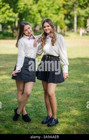 Zwei fröhliche Schüler in koordinierter weißer Kleidung und Fliege posieren zusammen auf einem grünen Feld und markieren den Beginn ihres Schuljahres Stockfoto