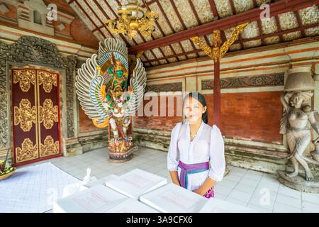 Bali, Indonesien - 11. November 2024: Junge Frau in traditionellem weißem Kebaya und farbenfroher Schärpe besucht einen balinesischen Tempel mit leuchtend grünen Garuda s Stockfoto