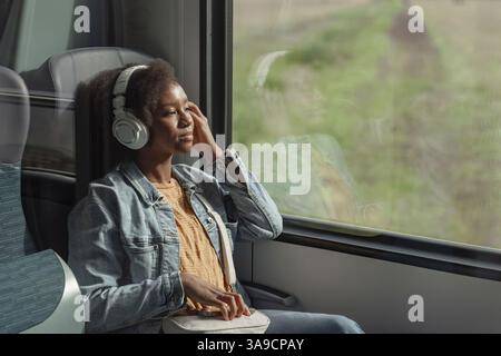 Junge afroamerikanische Frau sitzt bequem am Zugfenster, beobachtet die wunderschöne Landschaft und genießt Musik über ihre Kopfhörer. Stockfoto