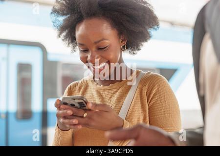 Lächelndes afroamerikanisches Reisenpaar mit Koffern, die auf einem Bahnsteig stehen, auf einen Zug warten und auf ein Handy schauen. Stockfoto