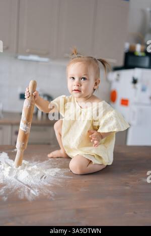 Vertikales Porträt eines fröhlichen Mädchens, das die Küche erforscht, mit Holznadeln und Mehl auf dem Tisch spielt, durch Spiel lernt und Stockfoto