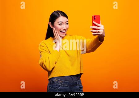 Wunderschöne asiatische Frau in einem Pullover, die ein Selfie mit ihrem Handy vor gelbem Hintergrund macht. Die junge Dame hält ihre Wange und lächelt lebendig, während sie ein Selbstporträt mit einem Mobilgerät macht. Stockfoto