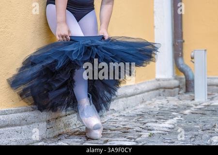 Schließen Sie junge weibliche Ballerina, die sich verkleidet und schwarzes Tutu an der gelben Gebäudewand anzieht. Ballett-Tänzer nimmt den Rock. Punkte Stockfoto
