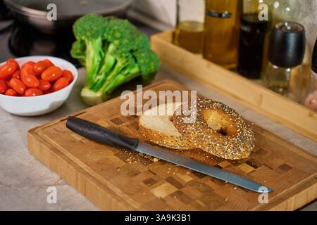 Geschnittener Bagel auf hölzernem Schneidebrett mit Kirschtomaten, Brokkoli und Küchenutensilien im Hintergrund. Zubereitung von gesundem Frühstück oder Snack zu Hause Stockfoto