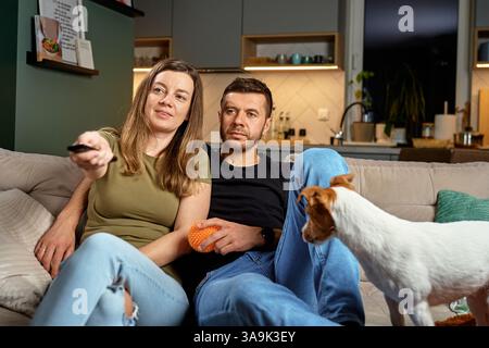 Mann und Frau sitzen auf Couch mit Fernbedienung und Hund. Paare schauen zusammen zu Hause fern. Konzept der häuslichen Entspannung, Gesellschaft und gemütliches häusliches Leben Stockfoto