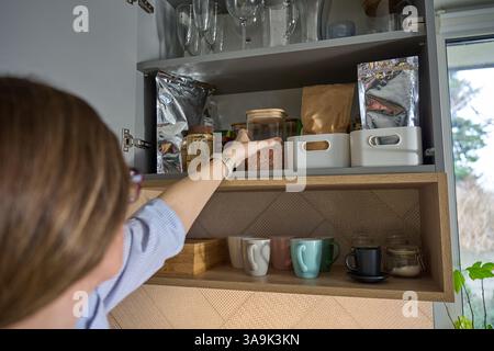 Die Person nimmt ein Glas aus dem Küchenregal, das mit Lebensmittelbehältern und Glaswaren gefüllt ist. Frau öffnet Schranktür im Küchenschrank Stockfoto