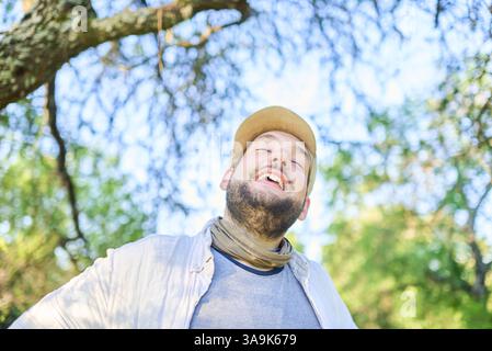 Porträt eines jungen Mannes, der mit einem Ausdruck von Glück und Wohlbefinden im Freien, in der Natur lächelt; der Hintergrund mit dem Kopierraum ist Himmel und Baum Stockfoto