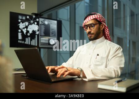 Ernsthafter junger Geschäftsmann aus dem Nahen Osten, der traditionelle Kleidung mit Brille trägt und in modernen Büros am Laptop arbeitet Stockfoto