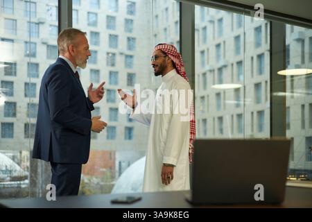 Zwei junge Geschäftsleute aus dem Nahen Osten und reife kaukasische Geschäftsleute, die im modernen Büro am Fenster stehen und über ein neues Projekt diskutieren Stockfoto