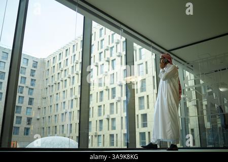 Weitwinkelaufnahme eines jungen Geschäftsmannes aus dem Nahen Osten, der aus dem Panoramafenster blickt, während er telefoniert, Kopierraum Stockfoto