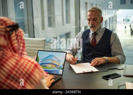 Überschulteraufnahme eines Reifen kaukasischen Geschäftsmannes, der vor einem Partner aus dem Nahen Osten sitzt und einen Vertrag mit dem Bauunternehmen unterzeichnet Stockfoto