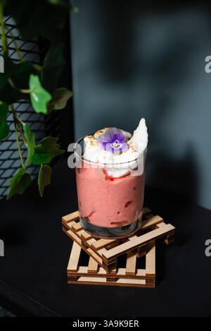 Ein Glas Erdbeermousse mit Schlagsahne und einer essbaren Blume, serviert auf einer hölzernen Achterbahn in stimmungsvoller Beleuchtung. Stockfoto