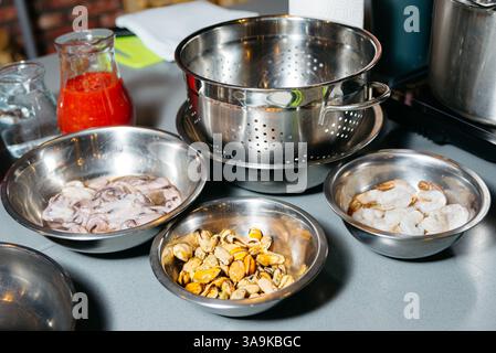 Eine Auswahl an frischen Meeresfrüchten, darunter Garnelen, Muscheln und Tintenfische, die in Metallschalen auf einer Küchentheke zum Kochen angeordnet sind. Stockfoto