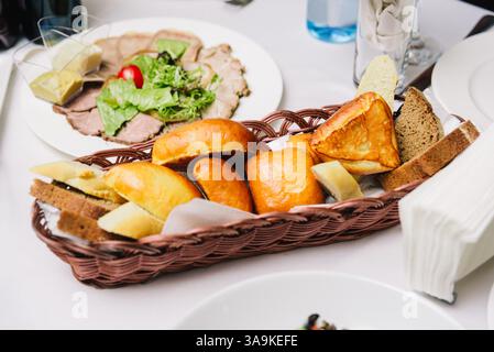 Verschiedene frisch gebackene Brotsorten und Gebäck, serviert in einem Korb auf einem weißen Tisch, ideal für eine Mahlzeit oder Vorspeise. Stockfoto