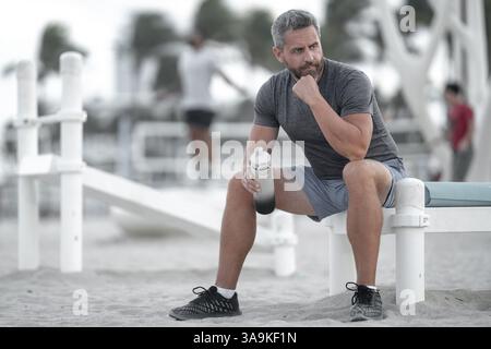 Sport-Fitness-Mann, der draußen auf einer Bank ruht. Reifes Fitnesssportmodell, das nach dem Training auf einer Bank sitzt. Ein Sportmann macht nach dem Fitnesstraining eine Pause Stockfoto