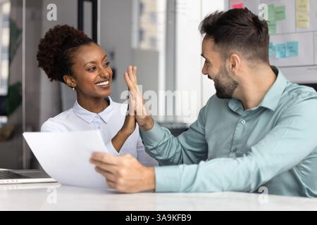 Ein paar glückliche, vielfältige Mitarbeiter, die am Tisch hohe fünf geben Stockfoto