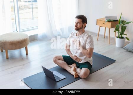 Mann, der vor dem Laptop auf dem Boden zu Hause meditiert Stockfoto