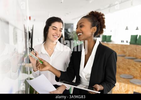 Zwei multiethnische weibliche Bürofreunde lachen über das Whiteboard Stockfoto